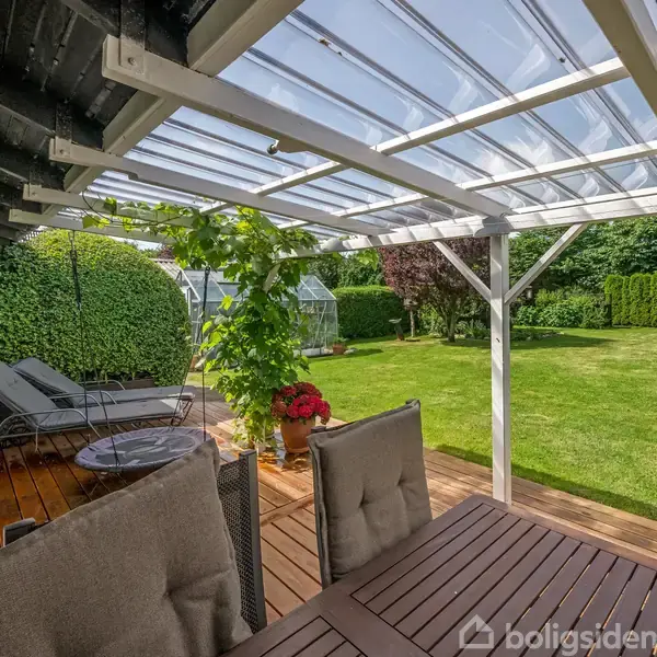 En træterrasse med spisebord og stole under et gennemsigtigt tag. I baggrunden er der en velplejet have med plæne, buskads og et drivhus.