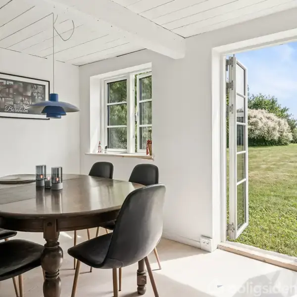 Et rundt spisebord med sorte stole omkring står i et lyst rum. Vinduer og åben dør viser udsigt til en grøn have med blå himmel. En plakat hænger på væggen.