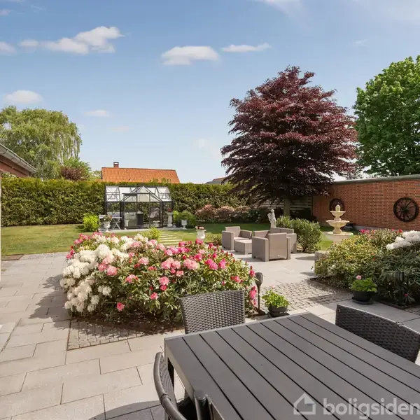 En terrasse med sorte havemøbler omgivet af blomstrende buske og en velplejet have. I baggrunden ses en stengrå skulptur og en glaspavillon. Store træer skaber skygge.