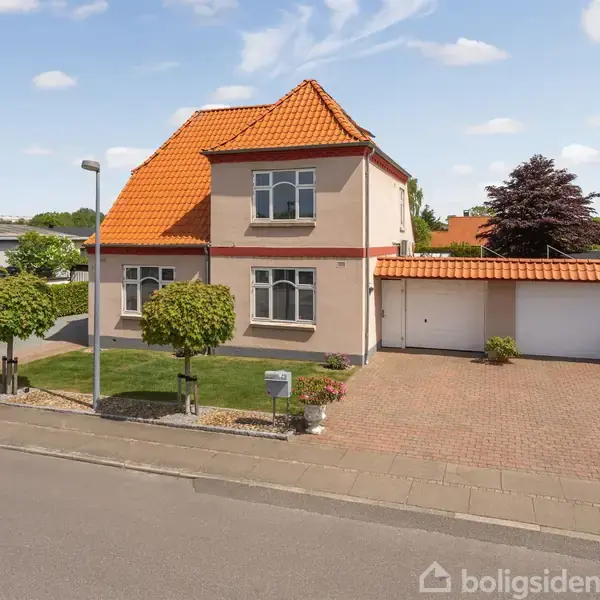 Et to-etagers hus med rødt tegltag står i en velplejet have med græsplæne og træer. En flisebelagt indkørsel leder til garagen. Flot dansk boligmiljø på en solrig dag.