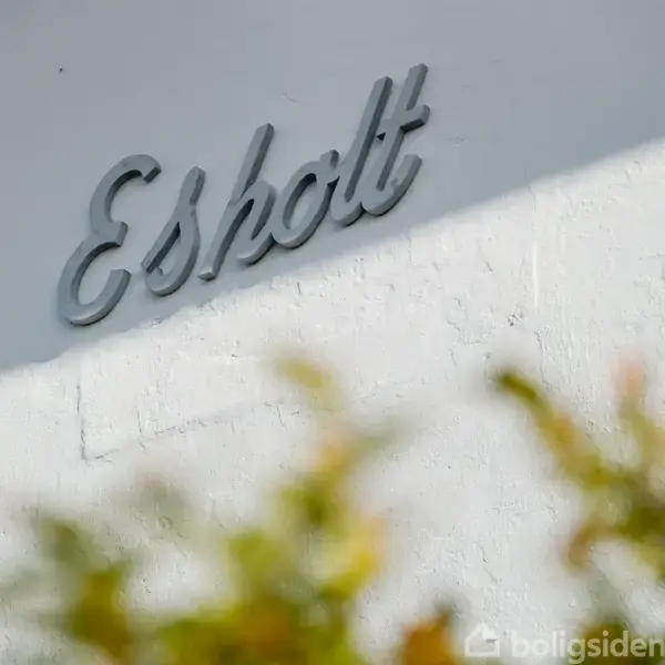 Skilt med teksten "Esholt" hænger på en hvid væg, omkranset af grønne og gule blade i forgrunden.