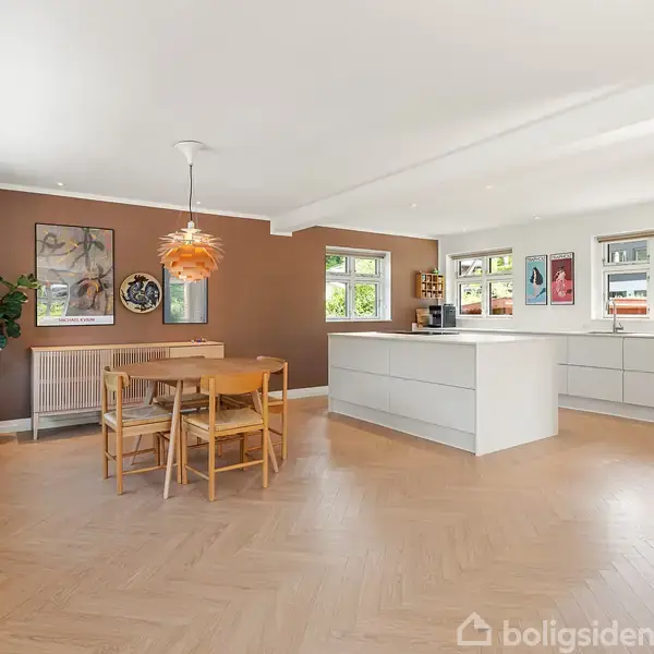 Et moderne køkken-alrum med hvidt køkken, køkkenø og spisebord. En orange lampe hænger over bordet. Væggene har kunstværker, og en plante står ved siden af radiatoren.