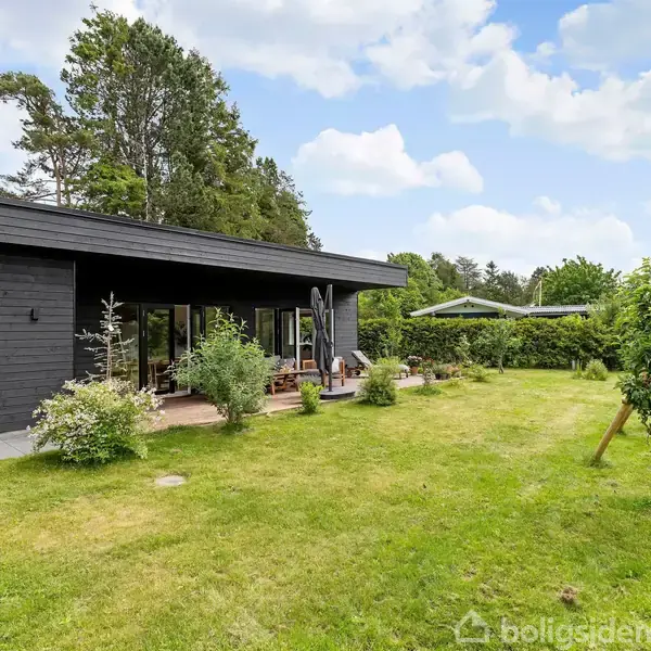 Sort, moderne hus med store vinduer, omgivet af en grøn græsplæne og buske. Terrasse med havemøbler til højre. Træer og blå himmel i baggrunden.