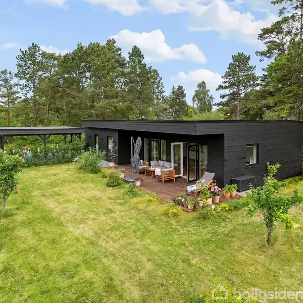 Et moderne sort hus står på en grøn græsplæne med et trædække i baggrunden. En træterrasse med møbler ses foran huset, omgivet af blomsterkrukker og en lille legehytte.