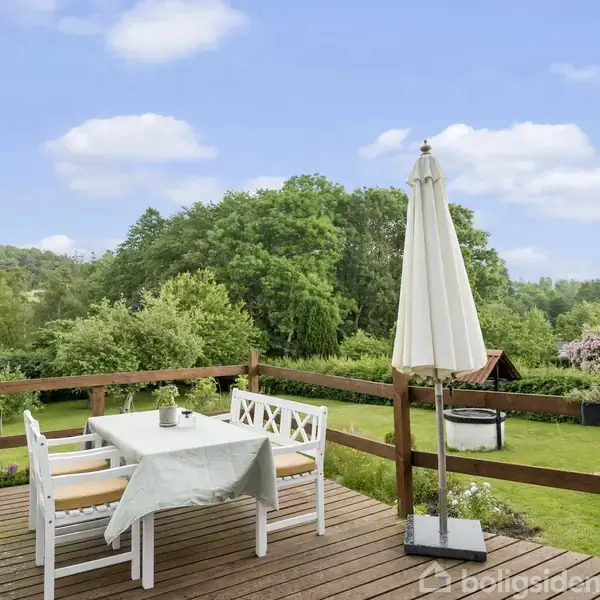 Et havesæt står på en træterrasse med en parasol, omkranset af frodig natur og grønne træer. Borde og stole er dækket med en dug.