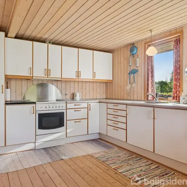 Et funktionelt køkken med hvide skabe og en komfur. En stribet løber dækker trægulvet. Et vindue til højre giver udsigt til en grøn have, omgivet af trævægge.