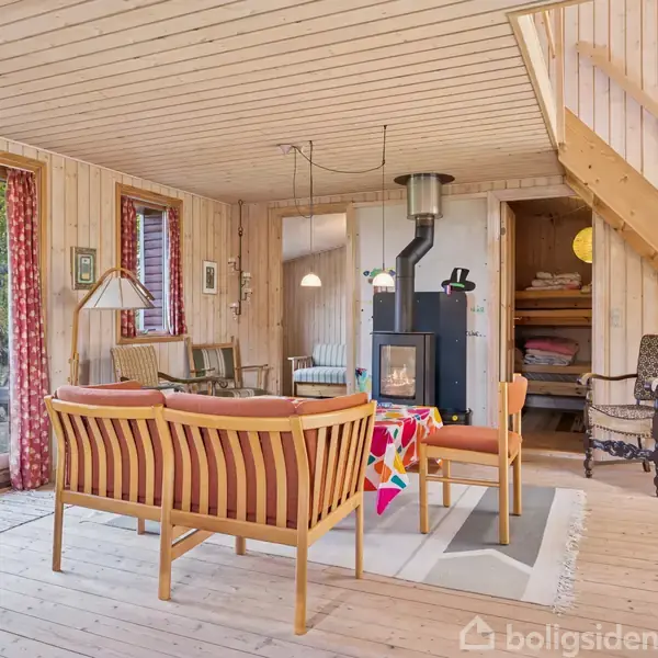 Stue med brændeovn i centrum, omgivet af trævægge og loft. Sofaer og stole står foran ovnen. Store vinduer giver udsigt til en grøn have.