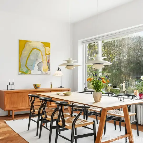 Et spisebord med seks stole står i en lys spisestue. Ved siden af hænger tre lamper over bordet. Et stort vindue viser en have udenfor. En abstrakt plakat pynter væggen.