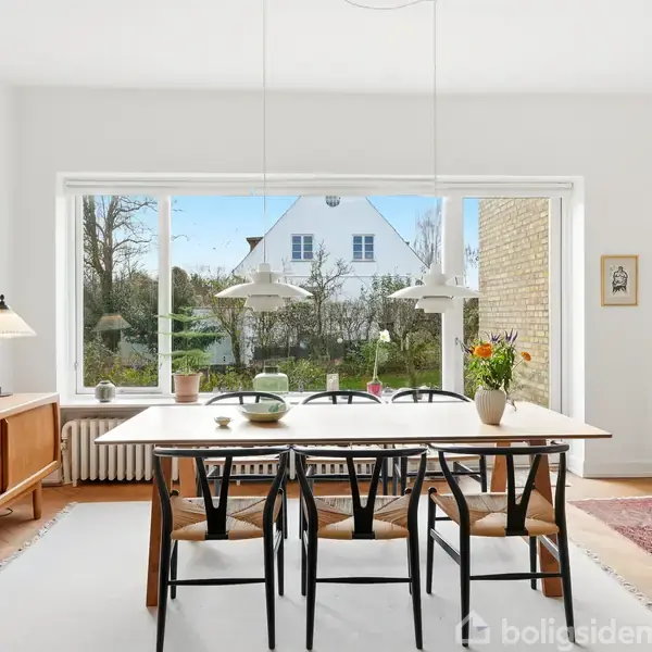 Spisebord med seks sorte stole står i et lyst rum ved et stort vindue. En skænk og lamper er bagved, og der er udsigt til en have og et hvidt hus.