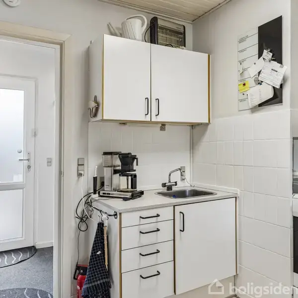 Et lille køkken med hvidt skab og vask. En kaffemaskine står på bordet. Der er en ovn til højre. Et bord står til venstre. Dør åbner til gang.