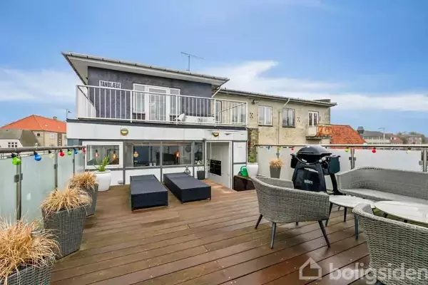 Træterrasse med loungemøbler, grill og planter. Ovenpå ses skiltet "Tandlæge" på en bygning med balkon. Omkringliggende huse i baggrunden under en blå himmel.