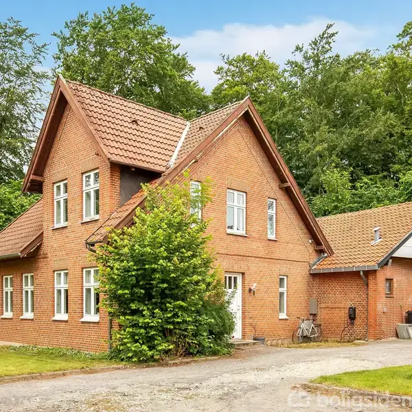 Et rødt murstenshus med skråt tag står omgivet af grønne træer og græsplæne. Ved siden af huset er en indkørsel og en lille carport. Der står en cykel ved husmuren.
