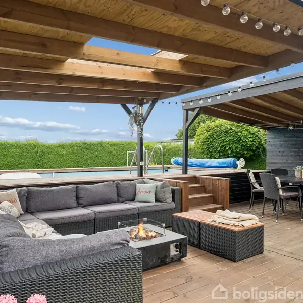 En udendørs terrasse med en stor hjørnesofa og bord, hvor der er opstillet en gaspejs. Foran er et spisebord med stole. Bagved ses en swimmingpool og grønne buske.