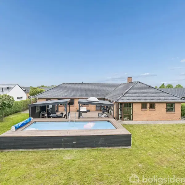 En moderne villa har en stor terrasse med en swimmingpool, omgivet af en velplejet græsplæne i et villakvarter. Huset har lyse murstensvægge og mørkt tag, under en klar himmel.