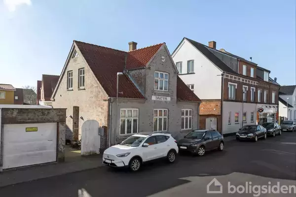 En hvid bil holder parkeret foran en grå murstensbygning med teksten "BAGETER." Bygningen ligger i en bymiljø med flere nabohuse langs en vej.