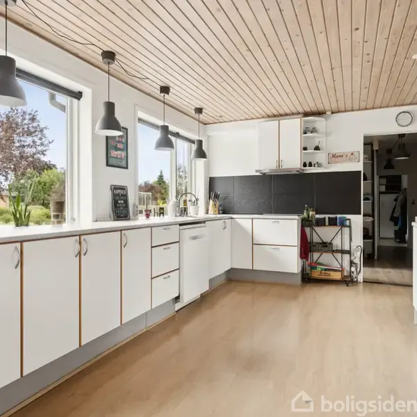 Køkken med hvide skabe og træloft. Tre hængende lamper oplyser rummet. Store vinduer giver udsigt til en grøn have. Døråbningen fører til en anden del af huset.