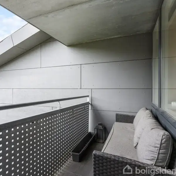 Balkon med en grå sofa, placeret mod en moderne facade. Omgivet af perforerede metalpaneler med udsigt til grøn vegetation. Klare glasruder afskærmer venstre side.