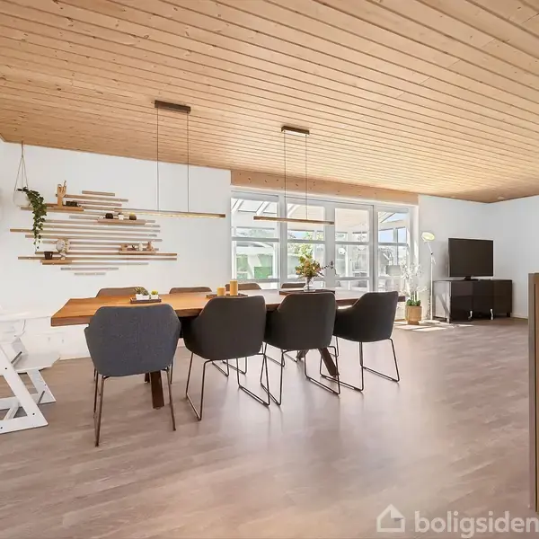 Spisebord med seks stole står i et moderne spisestueområde. Store vinduer tilføjer lys, mens træloftet tilføjer en hyggelig atmosfære. En tv-bænk med skærm står i baggrunden.