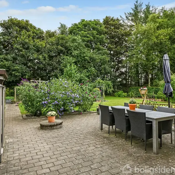 Terrassen er domineret af et bord med seks stole, hvor en parasol står ved siden af. Området er omgivet af frodig have, med blomsterbede og grønne træer i baggrunden.