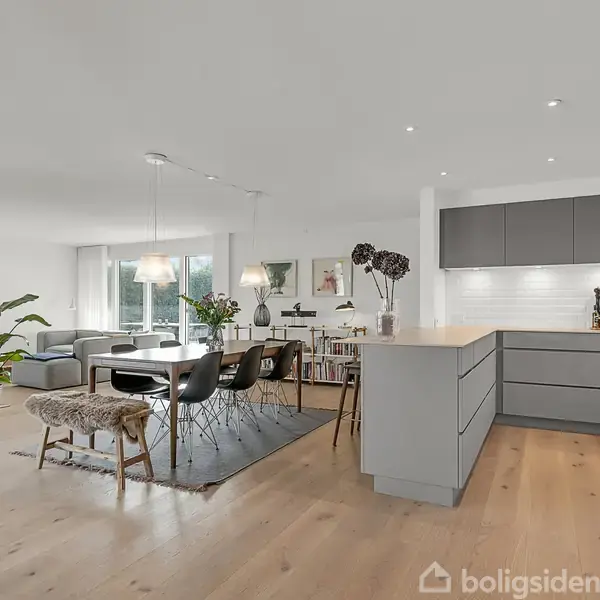 Køkken-alrum med moderne, grå køkkenskabe og spiseområde. Bordet er omgivet af sorte stole, og rummet har store vinduer og planter, hvilket skaber en lys og rummelig atmosfære.