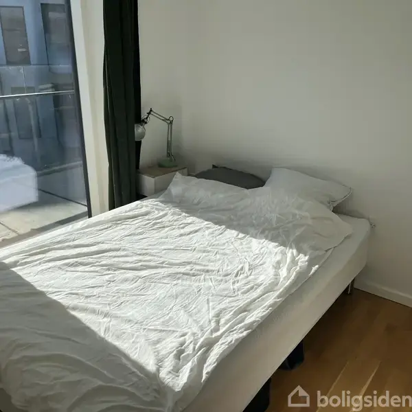 En seng med hvidt sengetøj står ved siden af en natbordslampe i et lyst værelse. Der er et vindue med mørke gardiner, der fører til en balkon.