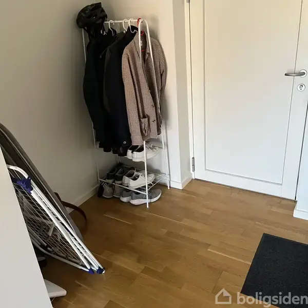 En række sko står på en skohylde ved siden af nogle jakker hængende på en tøjstativ. Tøjstativet er placeret i et hjørne af et rum med en hvid dør og trægulv.