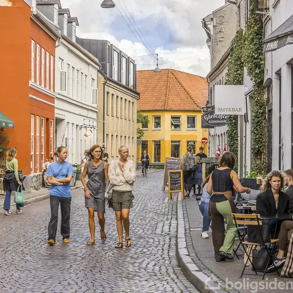 Folk går og sidder ved caféborde på en brostensbelagt gade med farverige bygninger. Skilte viser "Skyskraber Galleri" og "DRUKNENDE". Gaden har en livlig atmosfære med flere mennesker til stede.