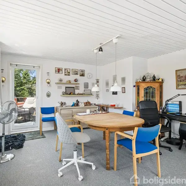Et rundt træbord står centralt med stole omkring. En computer står på et hjørneskrivebord. Vægge er dekoreret med billeder og genstande. En dør fører ud til en terrasse.