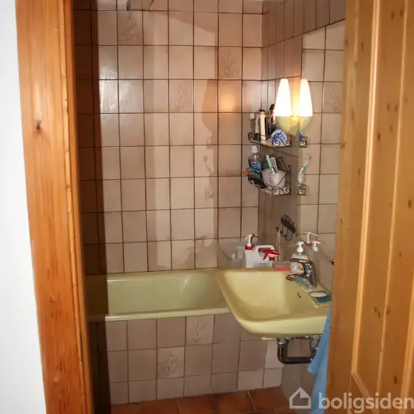 Badekar i en flisebelagt badeværelsesniche, omgivet af hylder med toiletartikler, oplyst af en væglampe. En håndvask er placeret ved siden af badekarret.