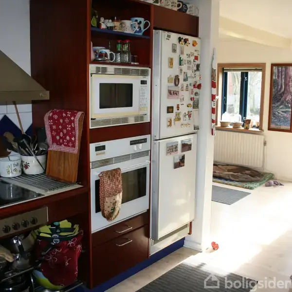 Køkken med komfur og emhætte til venstre, mikrobølgeovn og ovn i midten, køleskab med magneter til højre. I baggrunden ses spiseplads med vinduer, der giver naturligt lys.