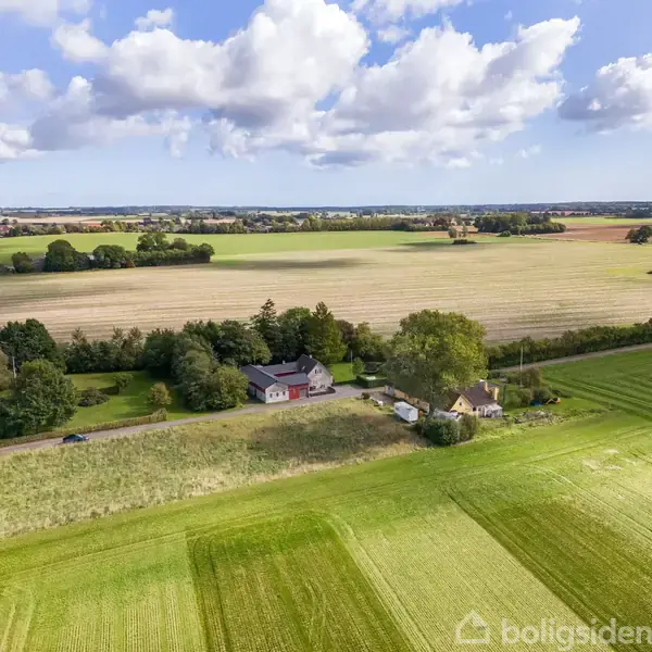 To huse ligger tæt på hinanden omgivet af grønne marker og træer under en blå himmel med spredte skyer. Landlig idyl med vidstrakt udsigt over det åbne landskab.
