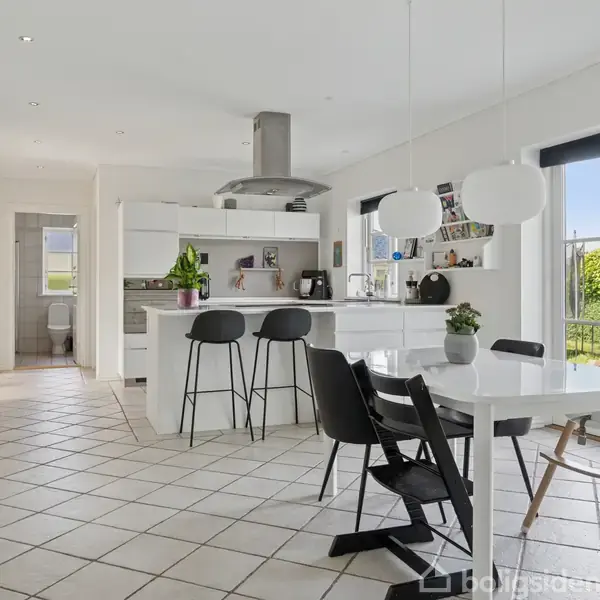 Et køkken med hvidt interiør har sorte barstole ved en køkkenø. I rummet er der et rundt spisebord med stole. Store vinduer giver udsigt til en grøn have.