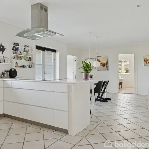 Et lyst, moderne køkken med en hvid ø, emhætte og åbne hylder. En plante står på bordet, sorte stole ved spisebordet, og åbent rum fører til andre værelser.