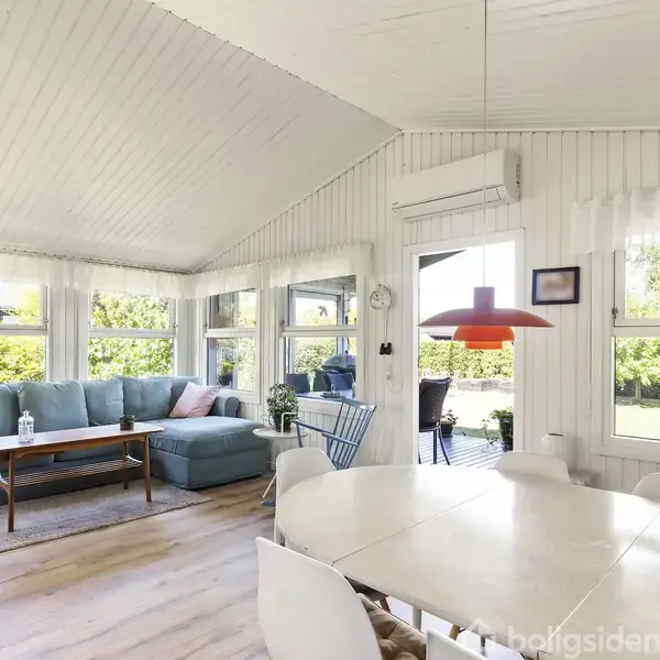 Stue med en hvid spisebord, blå sofa i hjørnet, og et træligende gulv. Store vinduer slipper naturligt lys ind, og en orange lampe hænger fra loftet. Gardiner pryder vinduerne.