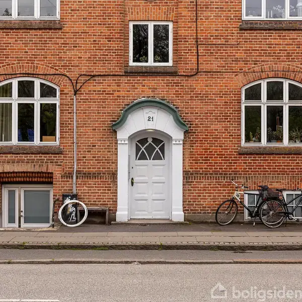 Rød murstensbygning med en centreret hvid dør markeret "21". Cykler står op ad muren. Vinduer flankerer døren, omkransende en urban gade.