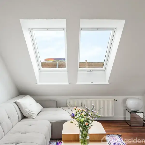 En lys stue med en grå sofa, sofabord med blomster og to skråvinduer ovenover. En radiator og et tv er placeret langs væggen, omgivet af hvide vægge.