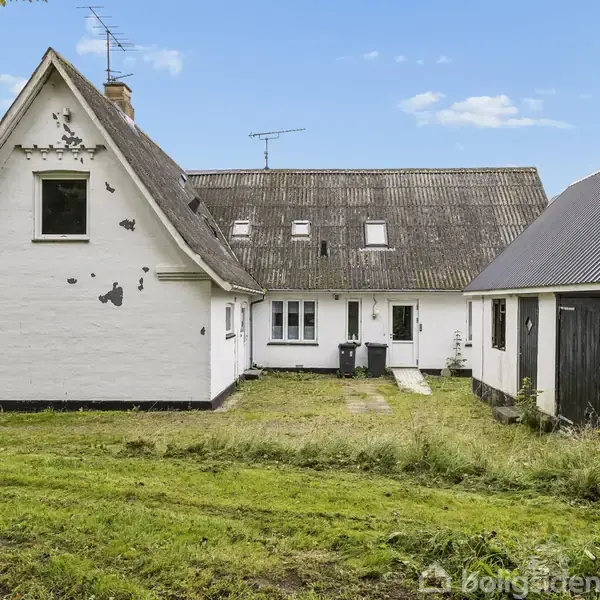 Et hvidt hus med skråt tag står stille i en have med græsplæne. Til højre ses en sort garagebygning. Himlen er blå med få skyer.