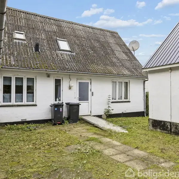 Hus med hvid facade og skråtag, der står stille i gårdhave med græs og fliser. Affaldsspande står ved indgangsdør. Ved siden af er der yderligere en bygning.