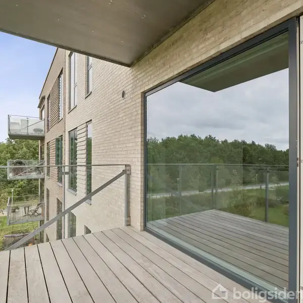En balkon med trægulv og glasrækværk åbner op mod et landskab med træer. Glasdøre står åbne ind til en moderne lejlighed med trægulv.