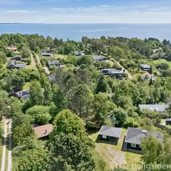 Flere huse ligger spredt blandt frodige grønne træer og buske, med udsigt over en blå kyst og havet i baggrunden. Stier snor sig gennem bebyggelsen.