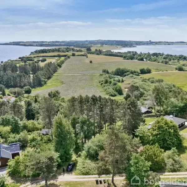 Huse ses fra oven omgivet af frodig skov og marker i et bakket landskab med udsigt over en fjord i baggrunden under en klar, blå himmel.