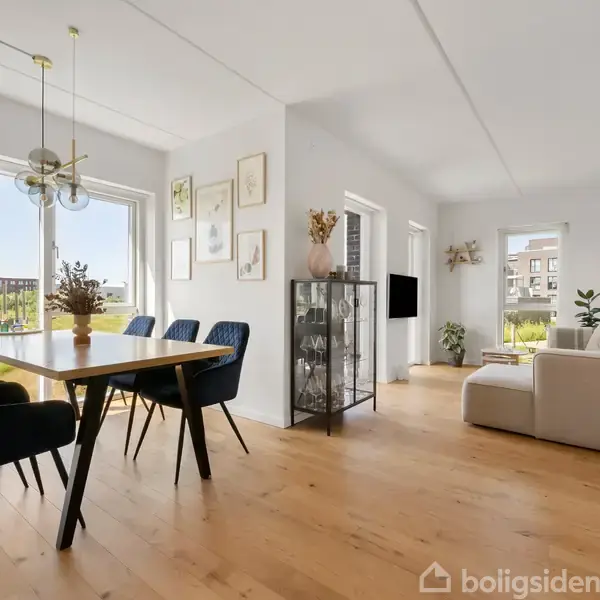 Et moderne køkken med hvide skabe og en indbygget ovn. En plante og krydderiglas står på bordpladen, mens gryder og beholdere hænger på en hylde. Trægulv skaber en varm atmosfære.