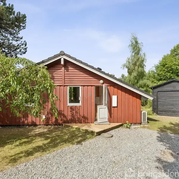 Rødt træhus står roligt i en naturskøn have omgivet af frodige træer og buske. Et grusbelagt stiareal fører op til huset og en sort trægarage står ved siden.