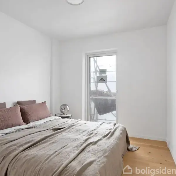 En seng er redt med beige sengetøj og brune puder i et lyst, minimalistisk soveværelse. Vindue i baggrunden med udsigt til en balkon eller lignende. Ingen tekst.