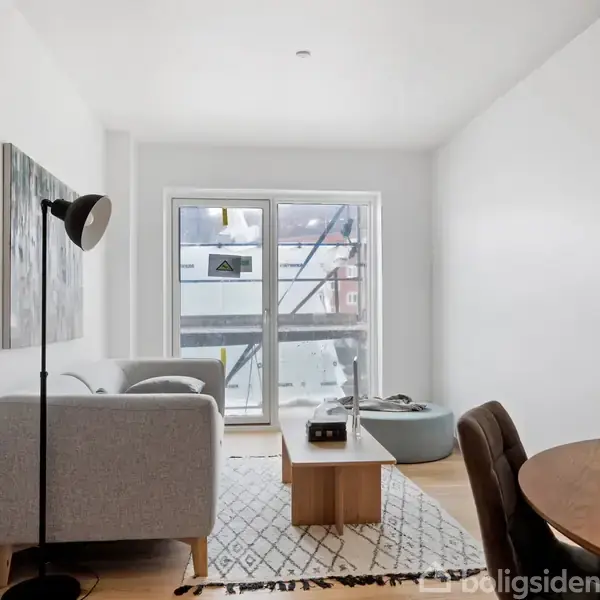 Grå sofa står i en lys stue ved siden af en gulvlapme; sofabord og spisebord er placeret på et lyst gulvtæppe. Vindue fuldender det minimalistiske rum.