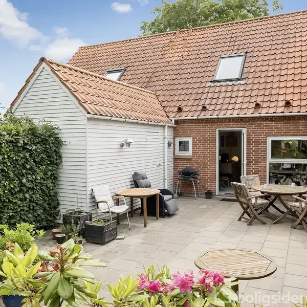 Hus med rødt tegltag og hvid træbeklædning står ved flisebelagt terrasse. Havemøbler og planter er placeret på terrassen, omkranset af grønne hække og under en blå himmel.