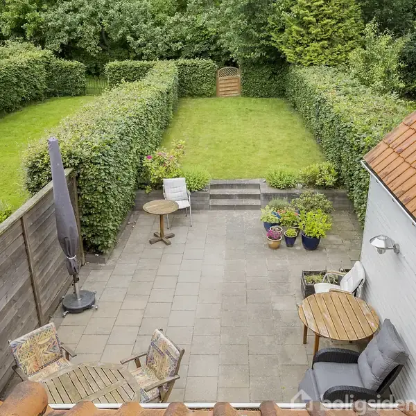 Terrasse med fliser og havemøbler. En parasol står ved siden af et bord og stole. Trappe fører op til en græsplæne omkranset af hække. Blomsterkrukker i hjørnet.
