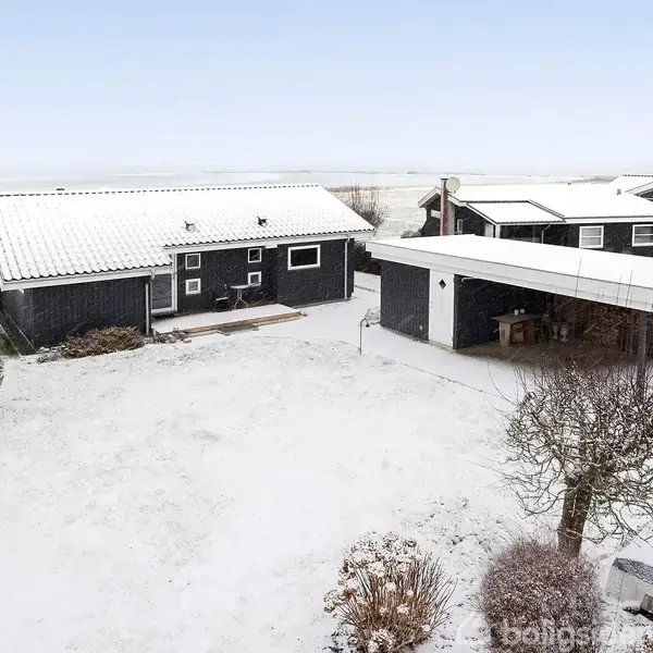 Et sort træhus står stille i en snedækket have ved siden af en carport. Sne hviler let på hustagene og de nøgne buske, mens landskabet forbliver fladt i baggrunden.