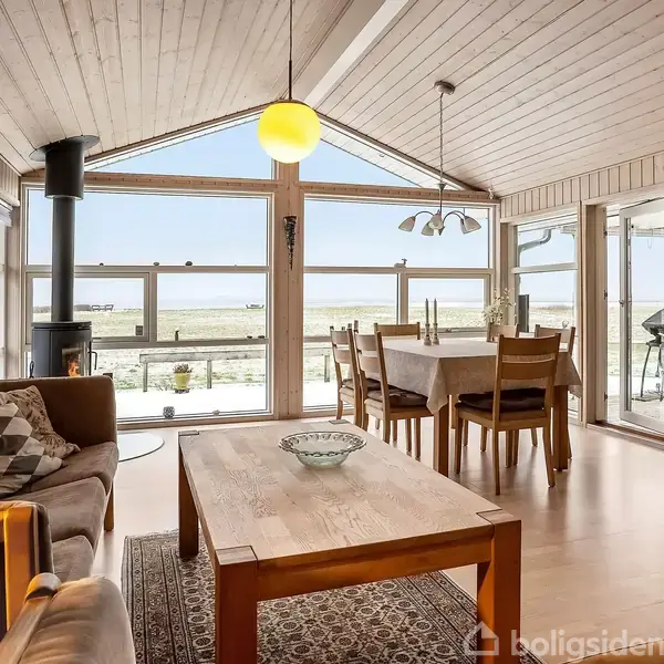 Stue med sofa og brændeovn, spisebord i træ ved panoramavinduer, udsigt over landskab. Lyse trævægge og loft, moderne indretning, lysindfald fra store vinduer, skaber en hyggelig atmosfære.