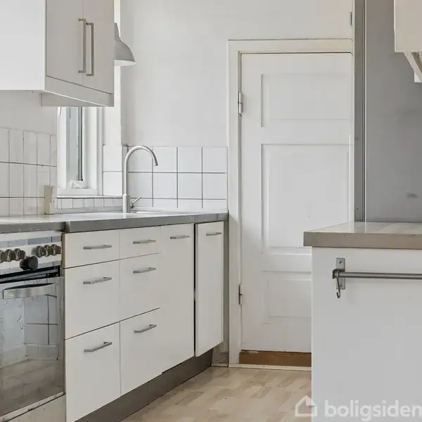 Køkken med hvide skabe og skuffer, en indbygget ovn, og en stålvask. Fliserne pryder væggen over bordpladen. En dør fører ud af køkkenet, og der er lyst trægulv.